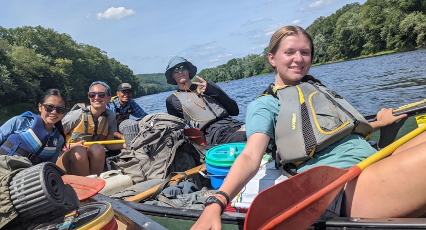 Several canoes holding two people each float near each other on a body of water framed by green trees. The people in the canoes are wearing life jackets and smile for the photo. 